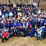 Large group of people gathered in forest setting, wearing casual and team shirts, posing for a group photo.
