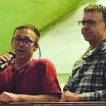 Two men speaking at an event, one holding a microphone, with a scenic landscape background.