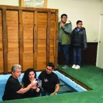People gathered for a baptism ceremony in an indoor pool, with others standing by to witness.