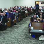 A large group gathered in a church hall, seated on pews, attentively listening to a presentation or sermon.