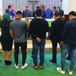 Group of people praying in a church, raising hands in prayer, dressed casually and formally.