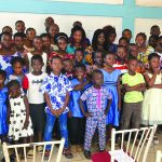 Group of diverse children and adults pose indoors, wearing colorful outfits and smiling at the camera.