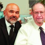 Two men dressed in a suit and tie, smiling at a social gathering with a blurred background.