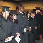Graduates in caps and gowns standing in a row during a ceremony, holding programs and showing solemn expressions.
