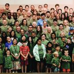 Group photo of diverse people wearing matching green event T-shirts, gathered inside a large room, smiling at the camera.