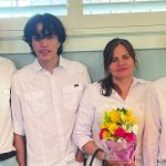 A family in matching white shirts poses indoors, smiling, with the woman holding a bouquet of colorful flowers.