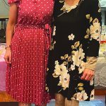 Two women in floral and polka dot dresses smiling at a gathering indoors.