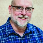 Portrait of a smiling man with glasses and a plaid shirt, standing against a patterned background.
