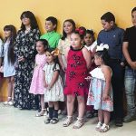A group of children and a woman stand against a yellow wall, dressed in colorful outfits, during an event or gathering.