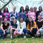 Group photo of diverse women outdoors, smiling together under trees on a grassy area.
