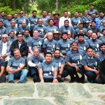 Group of men wearing matching shirts, posing outdoors in a park setting with trees and stone steps.