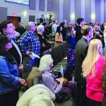 People gathered in a church service, standing and sitting, engaged in worship in a modern sanctuary.