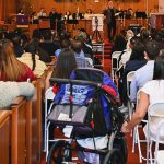 A diverse congregation attending a church service, viewed from the back, with stained glass windows and a choir in front.