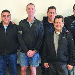 Group of ten men standing together indoors, smiling, casual attire.