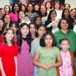 A diverse group of women and girls smiling at a community event, Vasijas de Honor banners in the background.