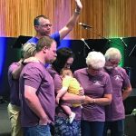 Group prayer gathering in a church setting, people in purple shirts supporting each other with a child.
