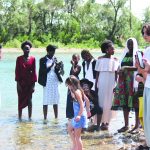 A diverse group of people enjoying a sunny day by the river, standing on the rocky shore and taking photos.