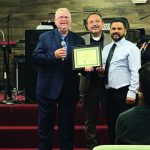 Group at a ceremony, man holding a certificate, smiling on a stage with musical equipment in the background.