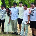 A group of friends stands closely together in a park, smiling under the shade of green trees.