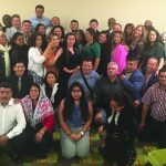Group of people at a gathering, posing and smiling in a room with a green carpet.