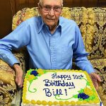 Elderly man celebrates 100th birthday with a decorated cake marked Happy 100th Birthday Bill while sitting on a sofa.