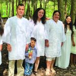 A group of people in white robes stands together in a wooded area, smiling.