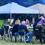 People enjoying a backyard gathering under tents with food, drinks, and conversation in an outdoor setting.