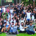 A large group of people joyfully raise their hands outdoors at Summit Lake Camp, surrounded by green trees and grass.