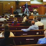People attending a church service, listening to a speaker in a wood-paneled sanctuary with red pews and floral arrangements.