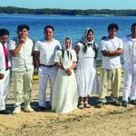 Group dressed in white standing on a beach by a lake, posing together with trees in the background under a clear sky.