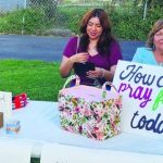 Two women at a table with a sign reading How can we pray for you today? offering prayer services.