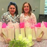 Two women smiling near a table with gift bags, high heels, and candles, in a cozy indoor setting.