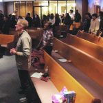 People standing in rows, engaged in a community gathering inside a hall with illuminated seating area.