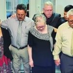 Group of people standing in prayer, holding hands in a room with closed eyes, conveying a solemn and united moment.