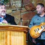 Man speaks at podium next to guitarist in church setting with stone wall background.