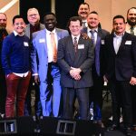 Group of men in formal attire smiling at an event, diverse in age and background, standing on a stage.