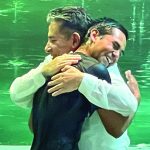 Two people hugging joyfully in front of a green backdrop, wearing white shirts, smiling broadly.