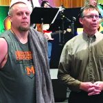 Two men standing together at an event, with musicians playing instruments in the background.