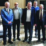 Seven men in business attire standing together indoors, smiling at the camera.