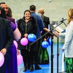 People holding pink and blue balloons, standing in line at an indoor event with microphones and a speaker.