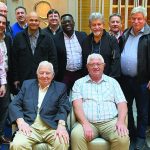 Group photo of 15 businessmen gathered indoors, two seated in front and others standing behind, smiling warmly.