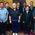 Group of ten diverse men standing indoors, posing for a photo in a warmly lit room with patterned wallpaper.