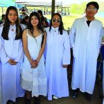 Group of people in white attire and one in a dress posing outdoors at an event with colorful tents in the background.
