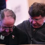 Two men in a moment of support, one comforting the other with a hand on his shoulder at an indoor event.