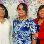 Three women posing in front of a floral backdrop, wearing elegant clothes: white, blue floral, and red jackets.