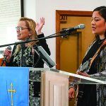Two women leading worship at a church podium with microphone and guitar.