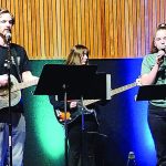 Musicians performing live with guitars and singing on stage in a vibrant setting with colorful lighting.