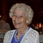 Elderly woman smiling at a social event, wearing a pearl necklace and white cardigan.