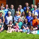 Large group of people posing outdoors in front of a cabin on a sunny day.
