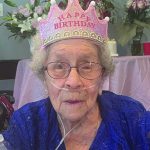 Elderly woman wearing a Happy Birthday crown celebrates her special day, surrounded by flowers and cake.